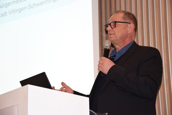 Ein Redner in Anzug und Brille steht am Pult, spricht ins Mikrofon und hält eine Präsentation.