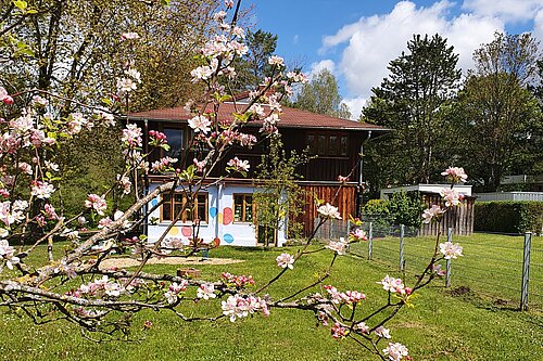 Zweistöckiges Holzhaus in großem Garten im Frühling; im Vordergrund blühende Obstbaumzweige, grüner Rasen und blauer Himmel.