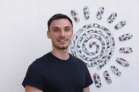 Junger Mann in schwarzem T‑Shirt lächelt vor einer weißen Wand, an der ein dekoratives Mosaik in Form einer Sonne mit spiralförmigem Kern hängt.