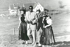 Schwarzweißfoto: Zwei Männer in Anzügen gehen Arm in Arm mit zwei Frauen in traditioneller Tracht über eine Wiese; im Hintergrund stehen Häuser. Die Gruppe wirkt fröhlich und vertraut.