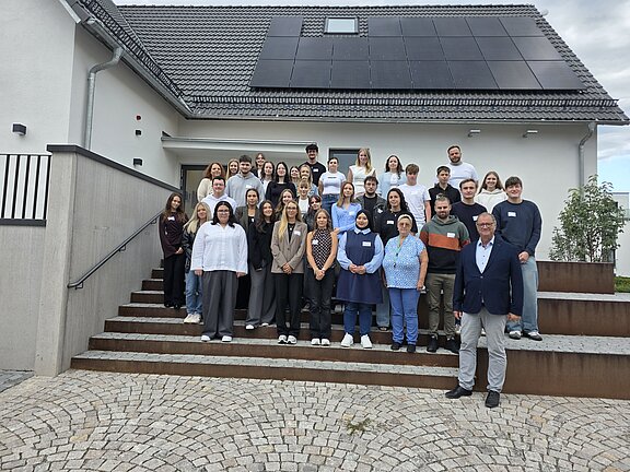 Gruppenfoto von etwa 35 Personen auf Außentreppen vor einem Haus mit Solarpaneelen auf dem Dach; Personen verschiedener Altersgruppen blicken zur Kamera.