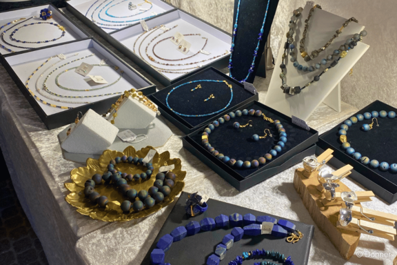 Auslage mit handgefertigten Schmuckstücken: mehrere Halsketten, Armbänder und Ohrringe aus Perlen und Steinen in Blau-, Grau- und Goldtönen, arrangiert auf schwarzen und weißen Präsentationskissen und Schalen.