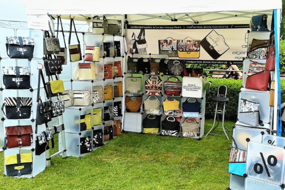 Marktstand unter Zelt mit zahlreichen Handtaschen und Umhängetaschen in verschiedenen Farben und Mustern, ordentlich auf Regalen und Haken ausgestellt, auf Grasboden.