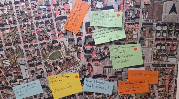 Luftbildkarte einer Stadt, beklebt mit mehreren bunten Notizzetteln, auf denen handschriftliche Anmerkungen und Pfeile zu Stadtbereichen zeigen, als Planungs- oder Beteiligungs-Tafel.