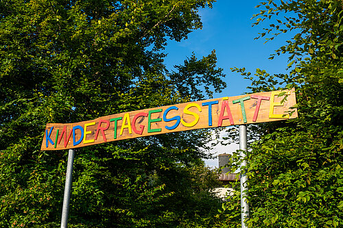Bunter Holzbalken mit der Aufschrift "KINDERTAGESSTÄTTE" als Eingangsschild, getragen von zwei Metallpfosten, vor grünem Baum- und Strauchhintergrund.