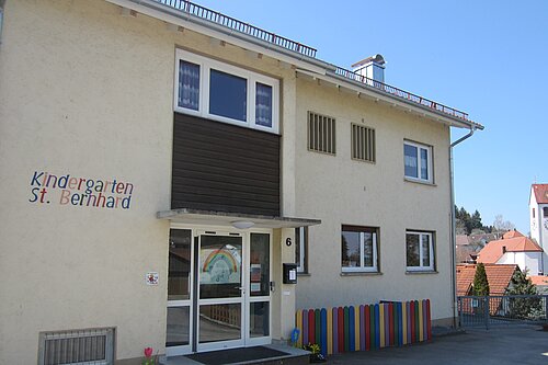 Eingang des Kindergartens St. Bernhard: helles zweistöckiges Gebäude mit bunter Zaunverkleidung und Regenbogenzeichnung an der Glastür; Kirchturm im Hintergrund.