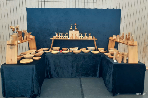 Marktstand mit auf dunklem Tuch arrangierten handgefertigten Holzschalen, Bechern, Löffeln und kleinen Holzfiguren sowie -kegeln als dekorative und nützliche Gegenstände.