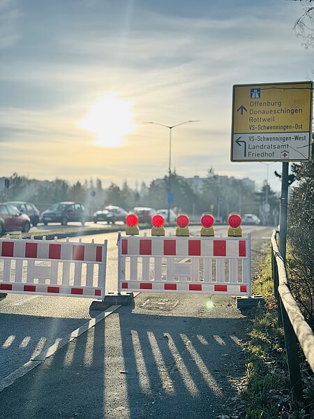 Baustellenabsperrung: rote-weiße Sperrbarriere mit roten Warnlichtern blockiert eine Straße; Richtungsschild weist auf Offenburg, Donaueschingen und Rottweil, niedrige Sonne am Horizont.