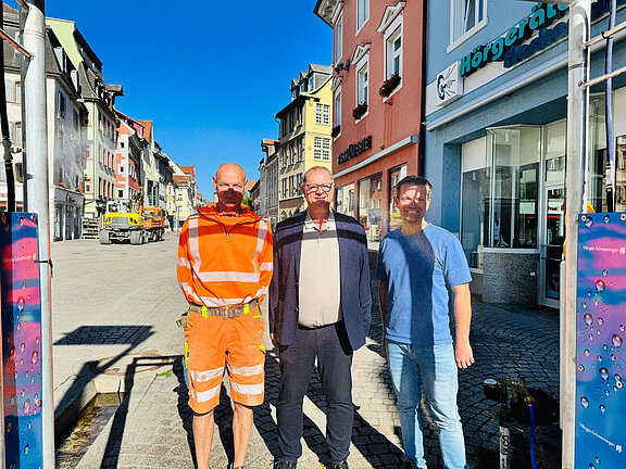 Drei Männer stehen nebeneinander auf einer sonnigen Fußgängerzone vor Geschäften; einer trägt orange Warnkleidung, einer ein Sakko, der dritte ein blaues T‑Shirt und Jeans.