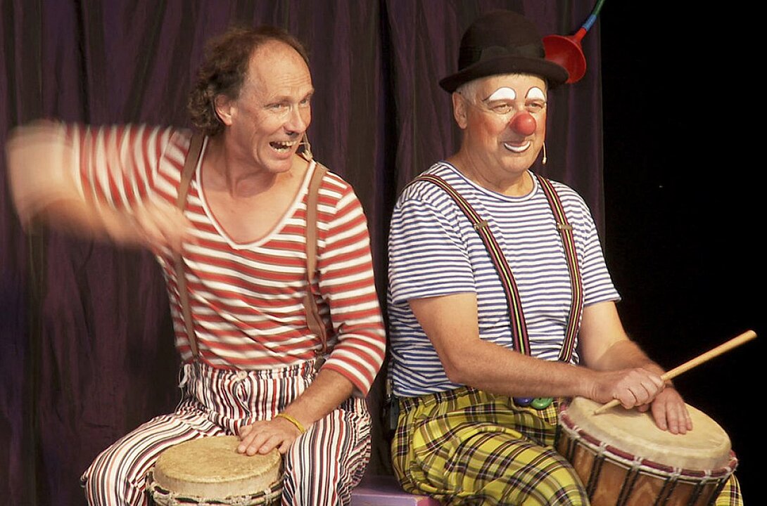 Zwei ältere Männer lachen und spielen Ukulele auf einer Bühne. (KI generiert)