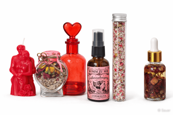 Anordnung kleiner Glasgefäße mit getrockneten Blüten, einem herzförmigen roten Flakon, einem Tropffläschchen mit Öl, einem Sprayfläschchen mit rosa Etikett und einer roten Kerze in Form eines umarmenden Paares.