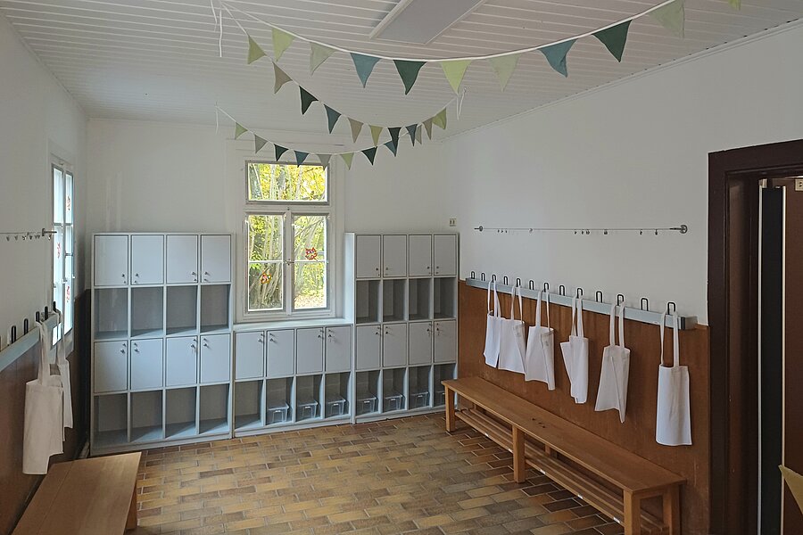 Heller Kindergarten-Garderobenraum mit niedrigen Schließfächern, Holzbänken, an Haken aufgehängten weißen Stofftaschen oder Schürzen und bunten Wimpelgirlanden an der Decke.