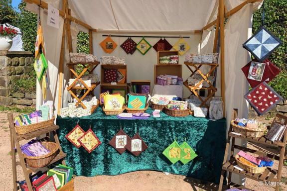Marktstand mit handgefertigten, bunten Stoff- und Patchworkartikeln: Topflappen, kleine Quilts, Stofftaschen und Karten, dekorativ auf Regalen, Tischen und hängend im Zelt präsentiert.