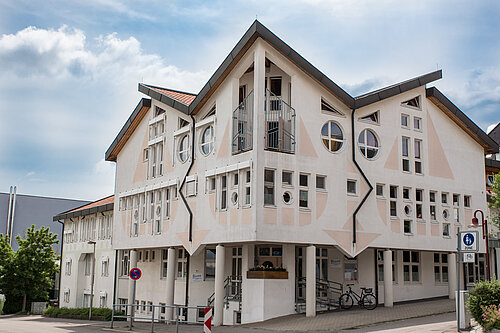 Dreigeschossiges weißes Gebäude mit spitzem Dach, markanten Giebeln, vielen runden und rechteckigen Fenstern, Säulen am überdachten Erdgeschoss und einem abgestellten Fahrrad an der Hausecke.