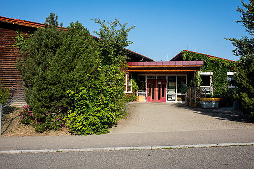 Eingang eines niedrigen, von Efeu und Sträuchern umwachsenen Gebäudes mit überdachtem Vordach und roten Doppeltüren; davor gepflasterte Zufahrt und sonniger blauer Himmel.