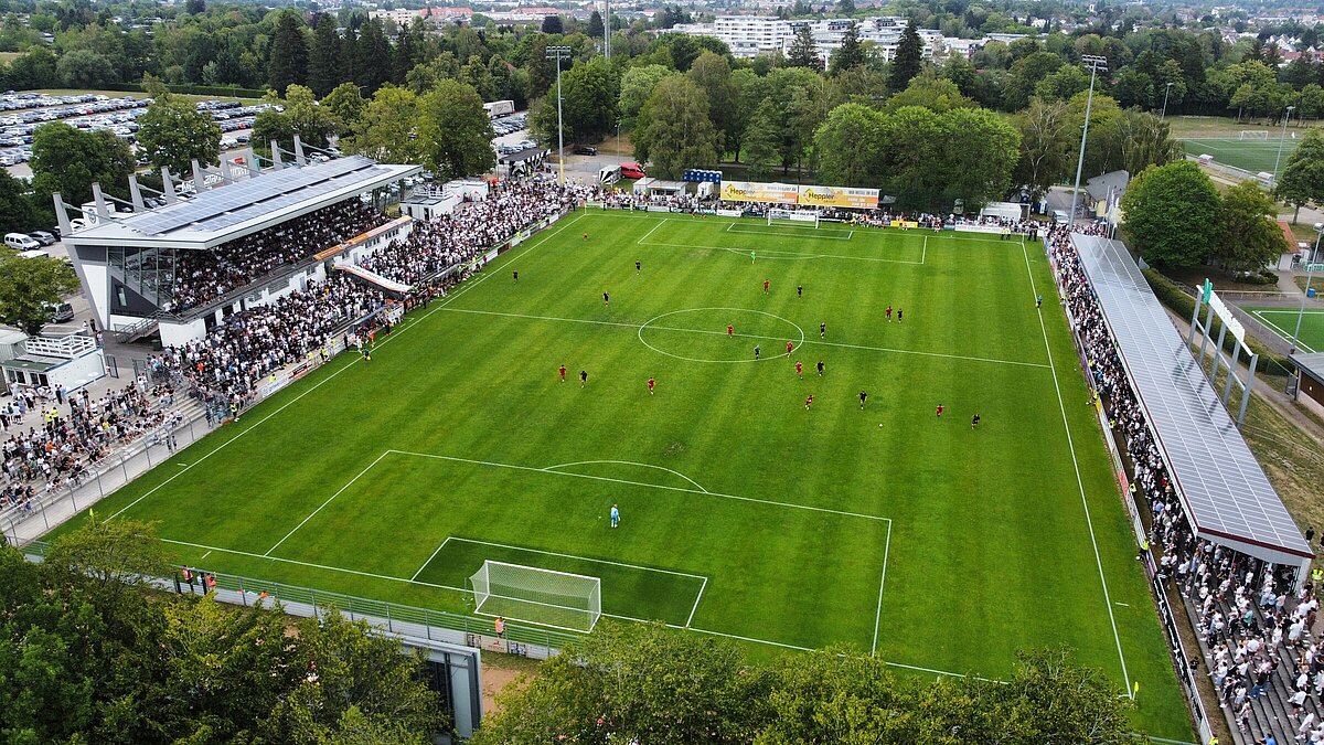 Luftaufnahme der MS Technologie-Arena während eines Heimspiels des FC 08 Villingen: grüner Rasen mit Spielern, voll besetzte Tribünen und Stehplätze