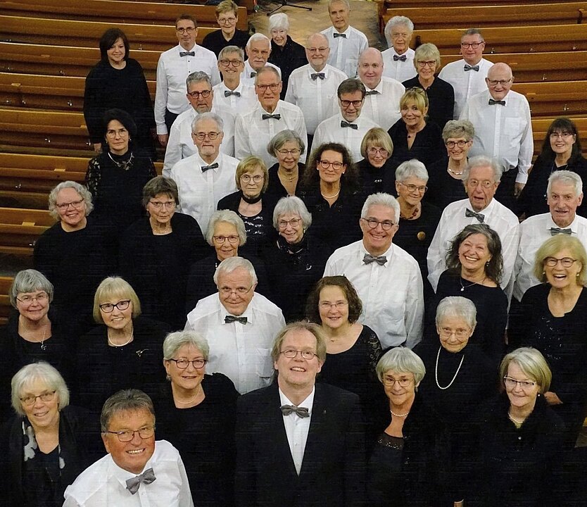 Gruppenfoto von 50 lächelnden Senioren in schwarzer Kleidung und weißen Hemden in einem Saal. (KI generiert)
