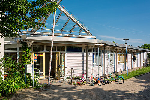Eingang eines niedrigen, hellen Gebäudes mit Glasvordach; vor dem Gebäude mehrere Kinderfahrräder und Roller aufgereiht, sonniger Tag.