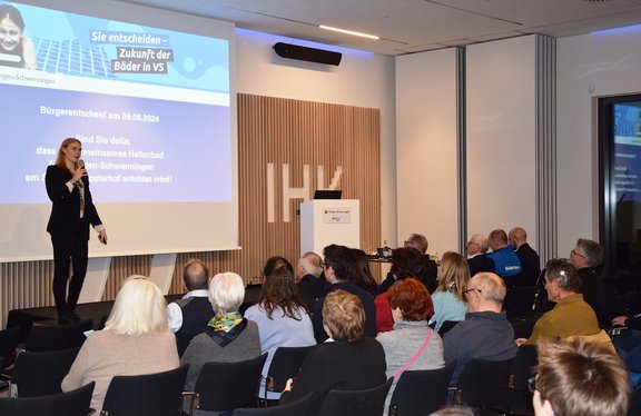 Referentin hält Vortrag über die Zukunft lokaler Bäder vor einem sitzenden Publikum in einem Konferenzraum; Projektionsfolie, Podium und aufmerksam zuhörende Menschen sind sichtbar.
