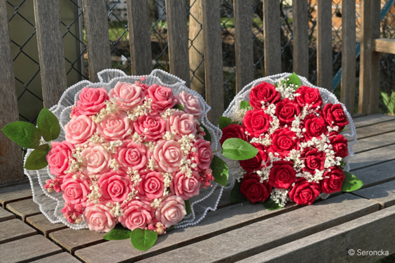 Zwei runde Rosensträuße auf einer Holzbank: links pastellrosa Rosen, rechts kräftig rote Rosen, beide mit weißem Schleierkraut, grünen Blättern und weißer Spitzenumrandung.