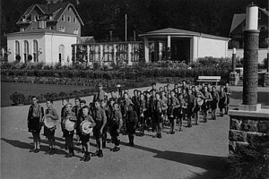 Schwarzweißfoto einer in Reih und Glied marschierenden Gruppe uniformierter Jungen, einige mit Trommeln, vor Gebäuden.