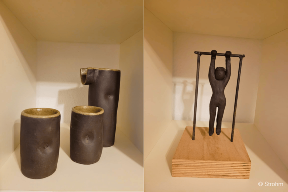 Regal mit drei dunklen Keramikgefäßen links (zwei Becher, ein hoher Krug); rechts eine kleine dunkle Skulptur einer hängenden Figur an einer Stange auf einem Holzsockel.