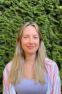 Porträt einer lächelnden Frau mit langen blonden Haaren, die ein lila Top und eine gestreifte Bluse trägt, vor einer dichten grünen Hecke.