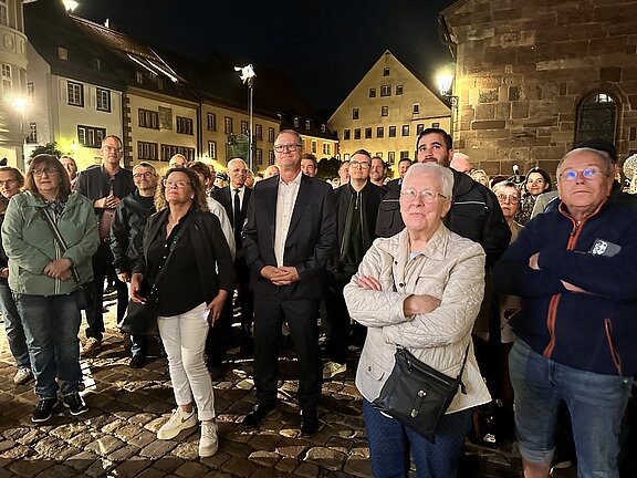 Menschenmenge in einem nächtlich beleuchteten historischen Marktplatz, mehrere Erwachsene stehen aufmerksam und erwartungsvoll, einige mit verschränkten Armen, Gebäude und Kopfsteinpflaster im Hintergrund.