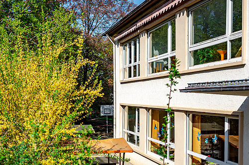Außenansicht eines zweistöckigen hellen Gebäudes mit großen Fenstern; davor eine Terrasse mit Tisch und Bänken, umgeben von grünen Sträuchern und blühenden Bäumen.