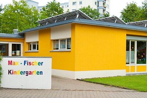 Gelb-orangener Kindergartenbau mit großen Glasfenstern, Eingangsbereich und Schild "Max‑Fischer Kindergarten" neben einer Rasenfläche.