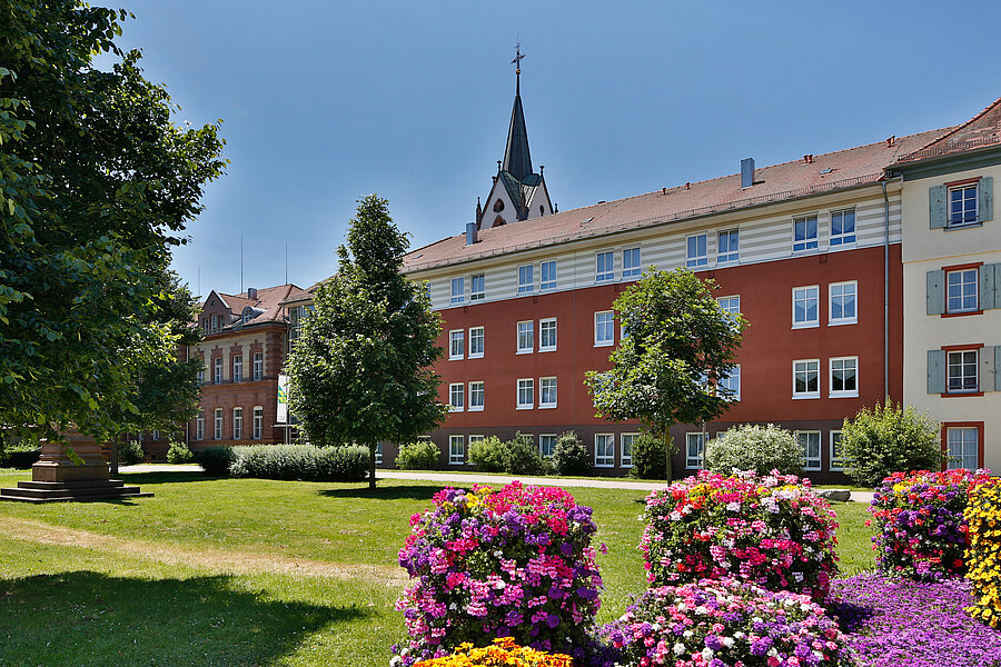Mehrstöckiges rotes Gebäude mit weiß gerahmten Fenstern, davor gepflegter Rasen, bunte Blumenbeete und Bäume; Kirchturm mit spitzem Turmhelm im Hintergrund.