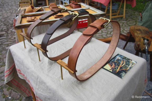 Drei breite handgefertigte Ledergürtel in verschiedenen Brauntönen mit Messing-Schnallen auf Holzständern präsentiert auf einem Markttisch.