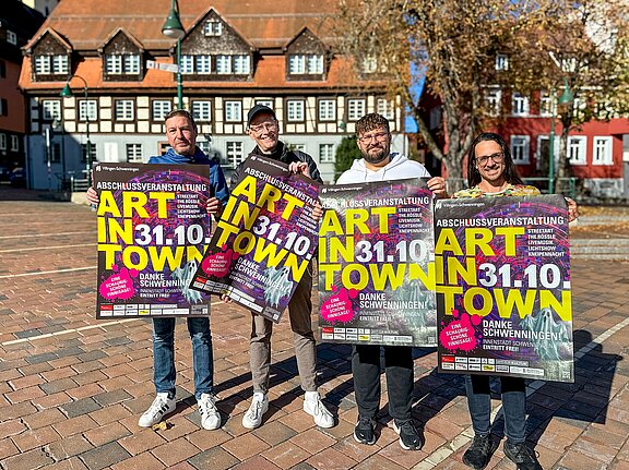 Vier Personen stehen auf einem Marktplatz und halten bunte Plakate mit der Aufschrift "ART IN TOWN 31.10", Werbung für eine Abschlussveranstaltung.