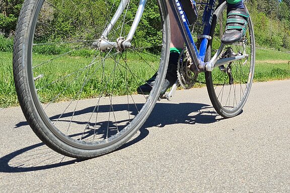 Nahaufnahme eines Fahrradfahrers in Sportkleidung auf einem Rennrad, der auf einer sonnigen Landstraße zwischen Bäumen und Wiesen fährt.