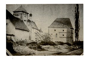 Schwarzweißaufnahme einer historischen Festungs- oder Klosteranlage mit massiven Steinbauten, quadratischem Turm, Kirche mit Zwiebelturm und unbefestigtem Vorplatz; rechts eine hohe Pappel.