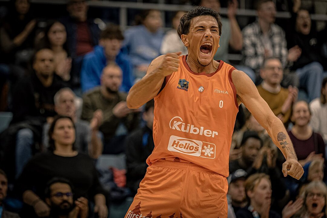 Jubelnder Sportler in orangefarbenem Trikot mit Sponsorenlogos, Hände hoch, Publikum im Hintergrund. (KI generiert)