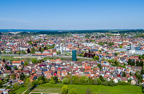 Luftaufnahme einer Kleinstadt mit roten Ziegeldächern, einem Fluss in der Bildmitte, Parks im Vordergrund und blauem Himmel.