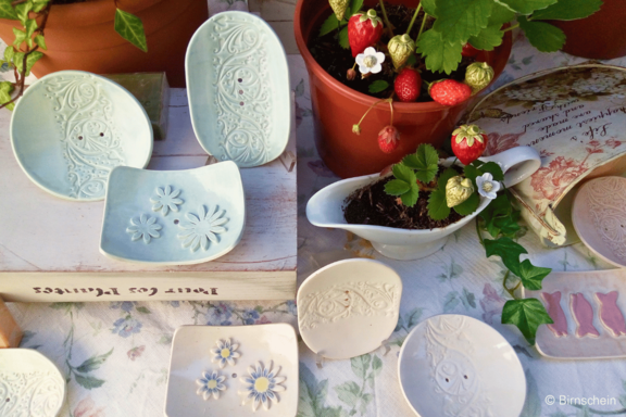 Handarrangement heller, handgearbeiteter Keramikschälchen und -seifenschalen mit floralem Relief, dekorativ aufgestellt neben einem Topf mit Erdbeerpflanze und roten Früchten.