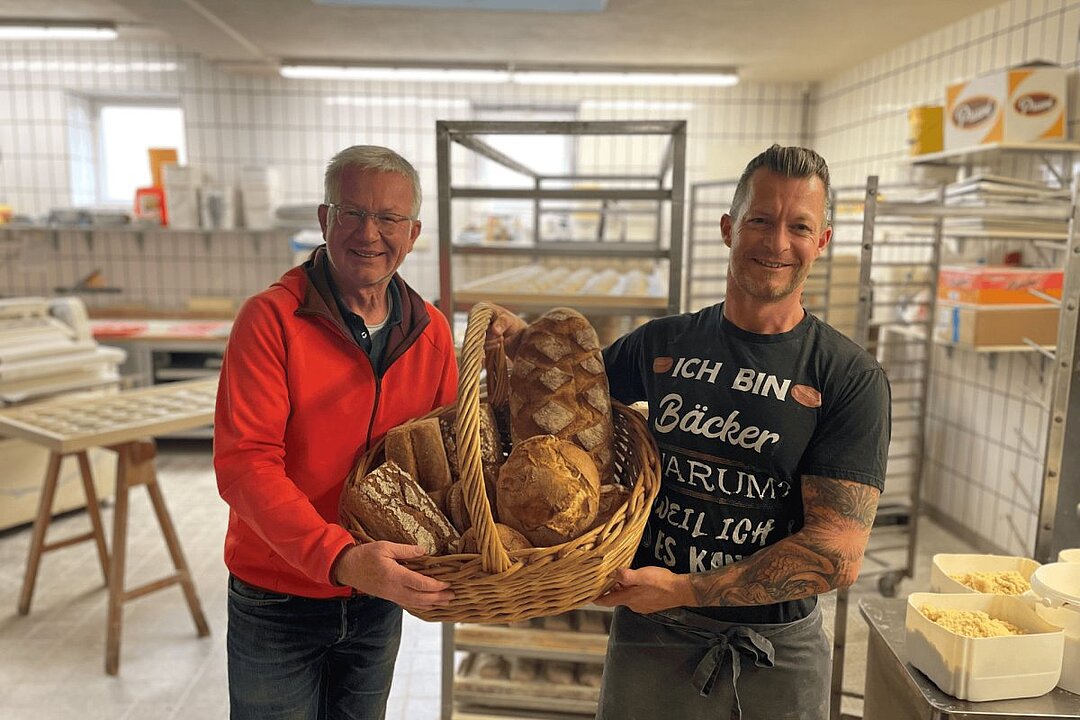 Zwei Männer in einer Bäckerei halten einen Korb mit Brot. Einer trägt ein T-Shirt mit der Aufschrift 'Ich bin Bäcker'. (KI generiert)