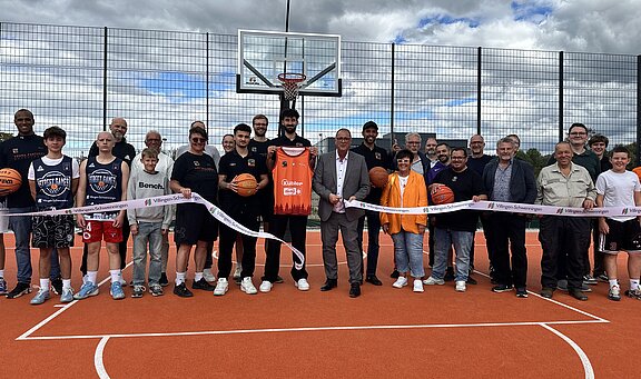 Gruppe von etwa 25 Menschen auf einem Außen-Basketballplatz vor dem Korb, posieren bei einer Eröffnungsfeier und halten ein Durchtrennungsband; einige tragen Basketballtrikots, eine Person präsentiert ein oranges Trikot.