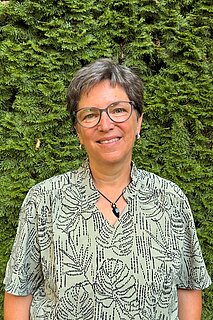 Porträt einer lächelnden Frau mittleren Alters mit kurzen grauen Haaren und Brille, in gemustertem hellgrünem Oberteil und schwarzem Anhänger, vor dichter grüner Hecke.