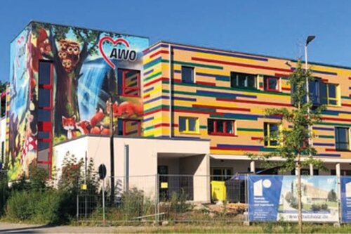 Buntes mehrstöckiges Gebäude mit farbiger Streifenfassade und großem Wandgemälde an der Ecke, AWO-Herzlogo sichtbar; umgeben von Bäumen und klarem blauem Himmel.