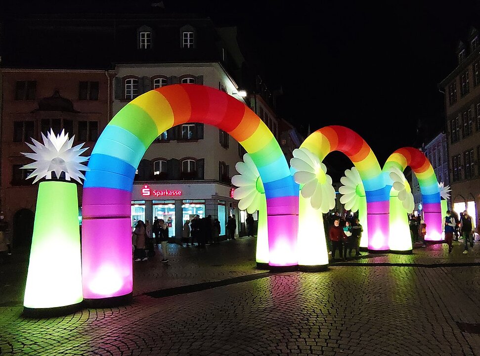 Bunte Luftballons in Regenbogenfarben vor einem historischen Gebäude bei Nacht. (KI generiert)