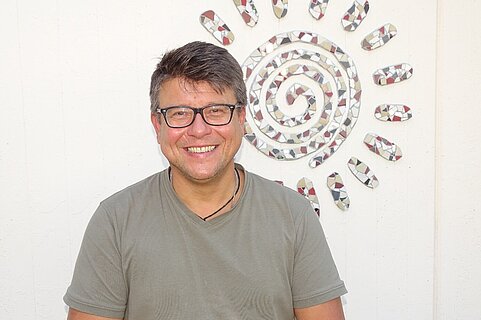 Lächelnder Mann mittleren Alters mit Brille und kurzem dunklem Haar, in grauem T‑Shirt, frontal zur Kamera vor weißer Wand mit dekorativem Mosaik-Sonnenmotiv.