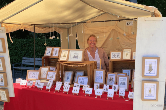 Lächelnde Verkäuferin in einem Marktstand, hinter einem roten Tisch mit zahlreichen gerahmten Illustrationen und kleinen Schmuckdisplayen.