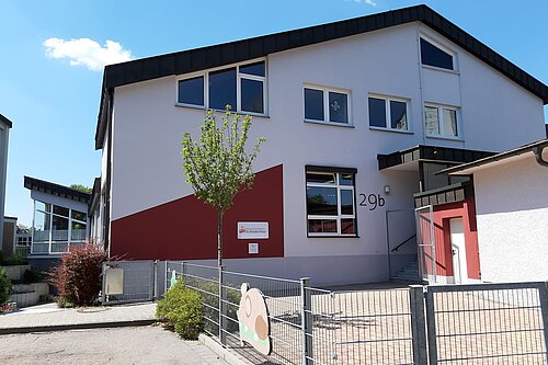 Zweistöckiges weißes Gebäude mit rotem Fassadendetail, Hausnummer "29b", Eingangsbereich, Metallzaun, kleinem Baum und spielerischer Dekoration am Zaun.