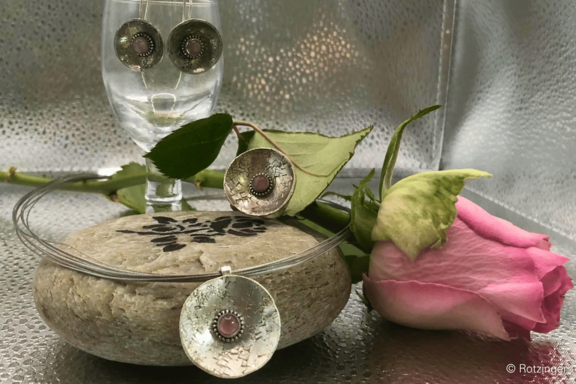 Silberschmuck-Set: rundes, gehämmertes Medaillon-Anhänger mit rosa Mittelstein und passende Ohrstecker, arrangiert auf einem runden Stein neben einer rosa Rose.