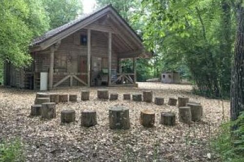 Rustikale Holzhütte im Wald mit Veranda; davor kreisförmig angeordnete Baumstumpf-Sitzblöcke auf Waldboden, wie ein offener Versammlungs- oder Lernplatz.