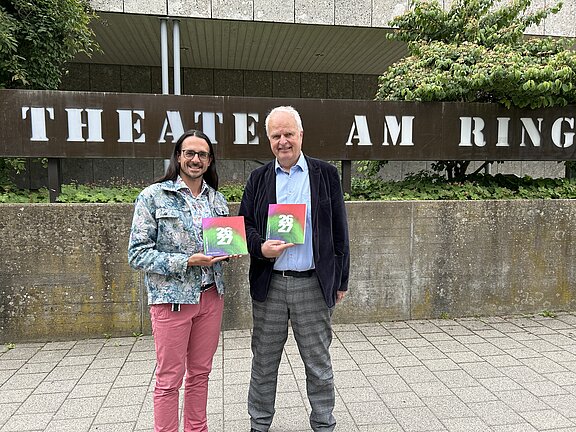 Zwei Personen stehen vor dem Schild „THEATER AM RING“ und halten jeweils ein buntes Programmheft; sie lächeln und posieren vor dem Theatereingang.