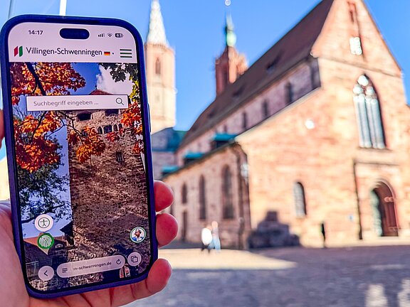 Ein Smartphone zeigt eine App mit einem Bild einer historischen Mauer, während im Hintergrund eine Kirche sichtbar ist.